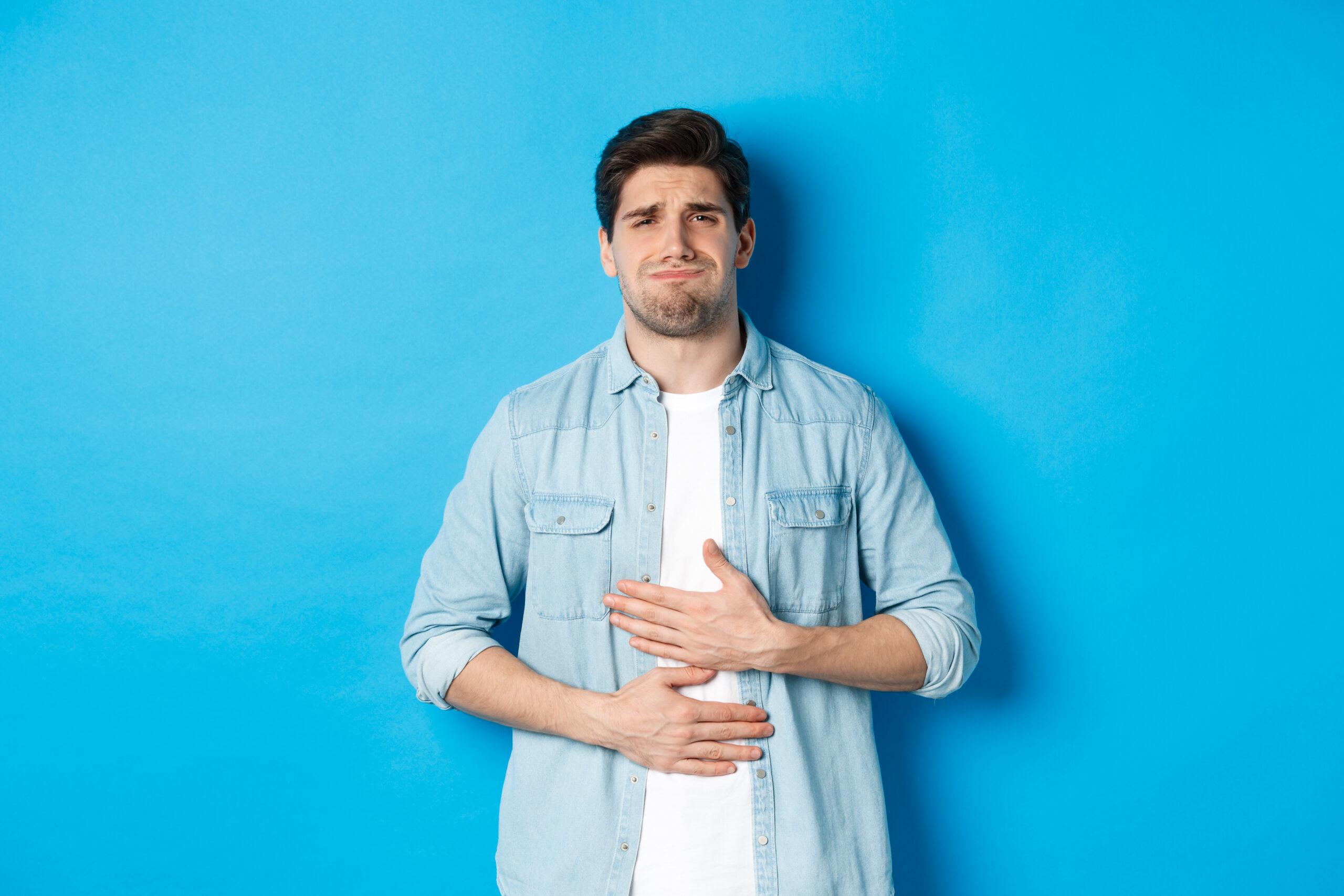man holding hands belly grimacing from pain complaining stomach ache standing against blue background scaled